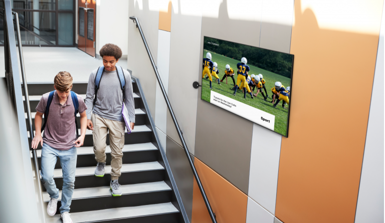 Two students walking down staircase next to digital sign displaying information about football game